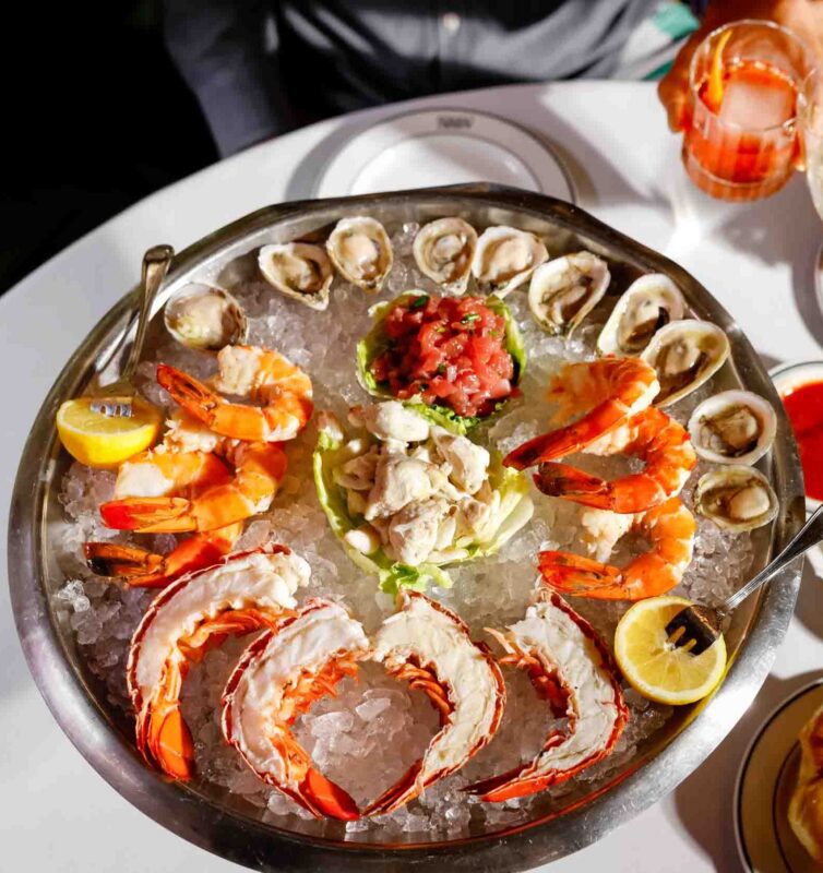 A platter of seafood on ice, featuring oysters, shrimp, lobster tails, and a tuna tartare center. Lemon wedges and cocktail sauce are included. A hand holds a drink in the background.