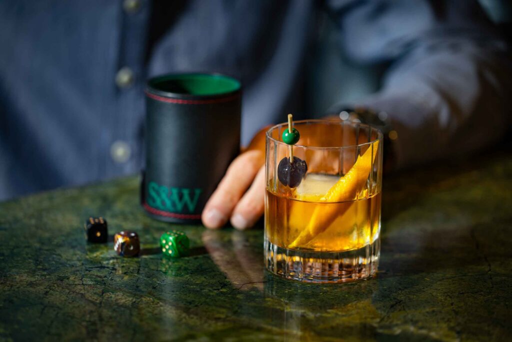 A person holds a black cup next to a cocktail with an orange peel and cherry garnish on a green marble table, with dice scattered nearby.