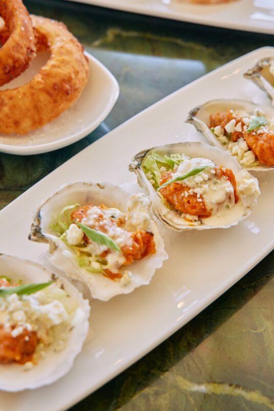 A row of oysters on the half shell topped with creamy sauce, green onions, and cheese on a white plate. Onion rings are visible in the background.