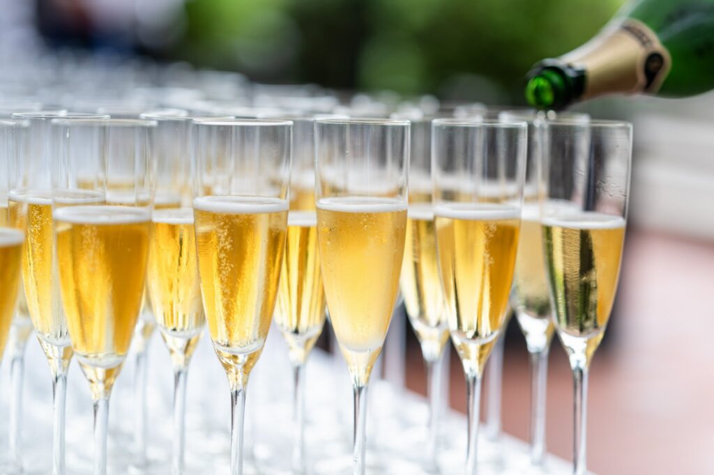 A close-up of several champagne flutes filled with sparkling wine, with one being poured from a green bottle, set on a white table outdoors.