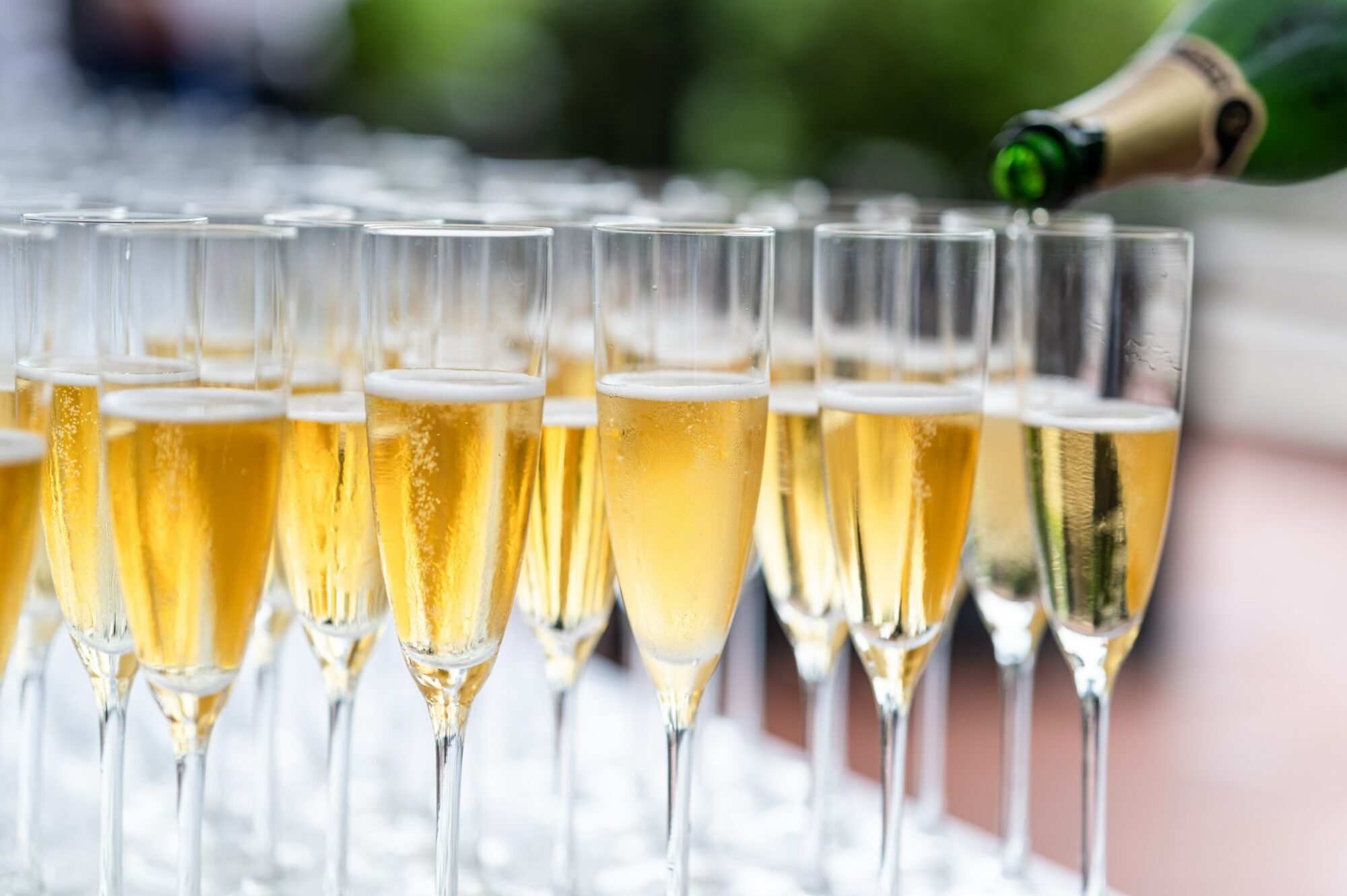 A close-up of several champagne flutes filled with sparkling wine, with one being poured from a green bottle, set on a white table outdoors.
