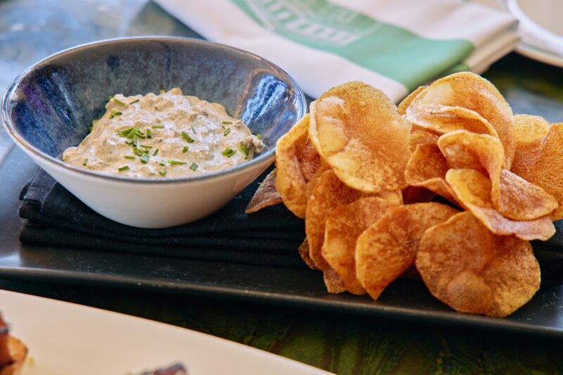 A bowl of creamy dip garnished with chopped chives sits next to a pile of crispy potato chips on a dark tray.