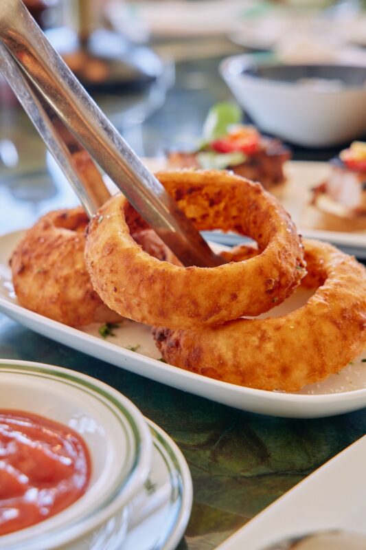 A pair of metal tongs holds a crispy onion ring above a plate stacked with more onion rings. A bowl of dipping sauce sits nearby on the table.