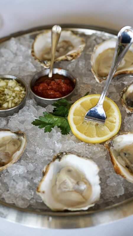 A round tray of raw oysters on ice, served with lemon slices, parsley, and small bowls of cocktail sauce and mignonette, with metal spoons.