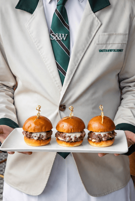 A server in a white jacket and green tie holds a rectangular plate with three cheeseburger sliders, each topped with melted cheese and a golden bun, secured with decorative picks—perfect bites from the Game Day Menu at Smith & Wollensky Atlantic Wharf for those looking to watch football in Boston.